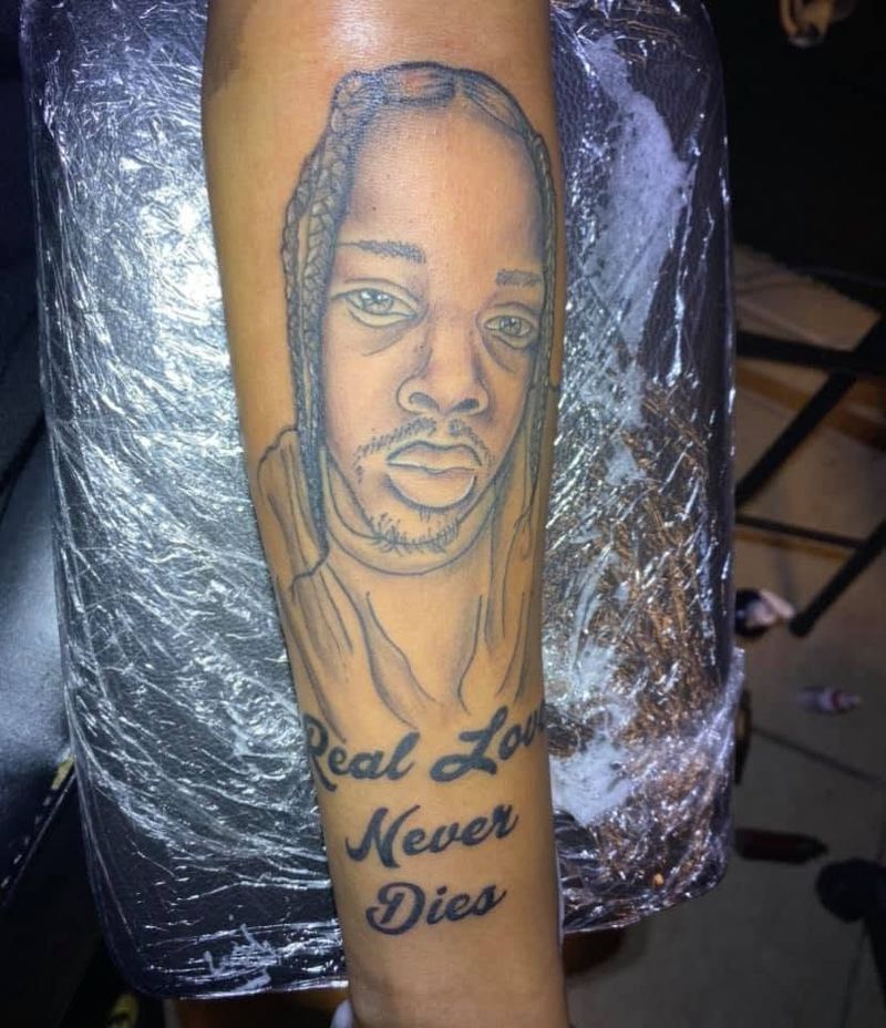 Black and grey portrait tattoo of a young man with braids above the script Real Love Never Dies on a forearm.
