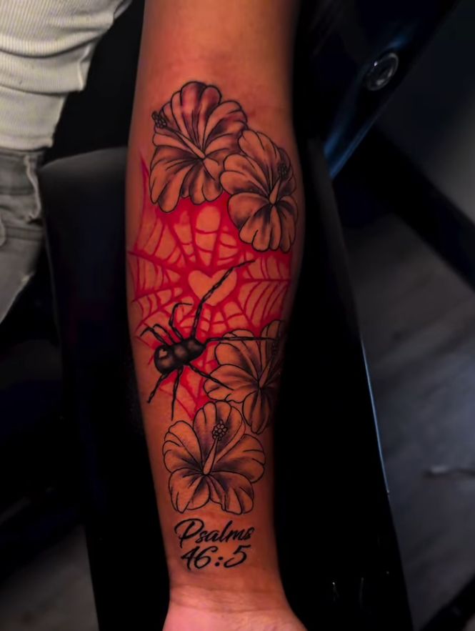 Color forearm tattoo with a red spiderweb, a black spider, orange flowers and script reading Psalms 46:5 near the wrist.